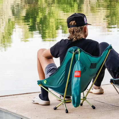 A man in a black shirt and cap sits in a black Grand Trunk Mantis chair by a calm lake.