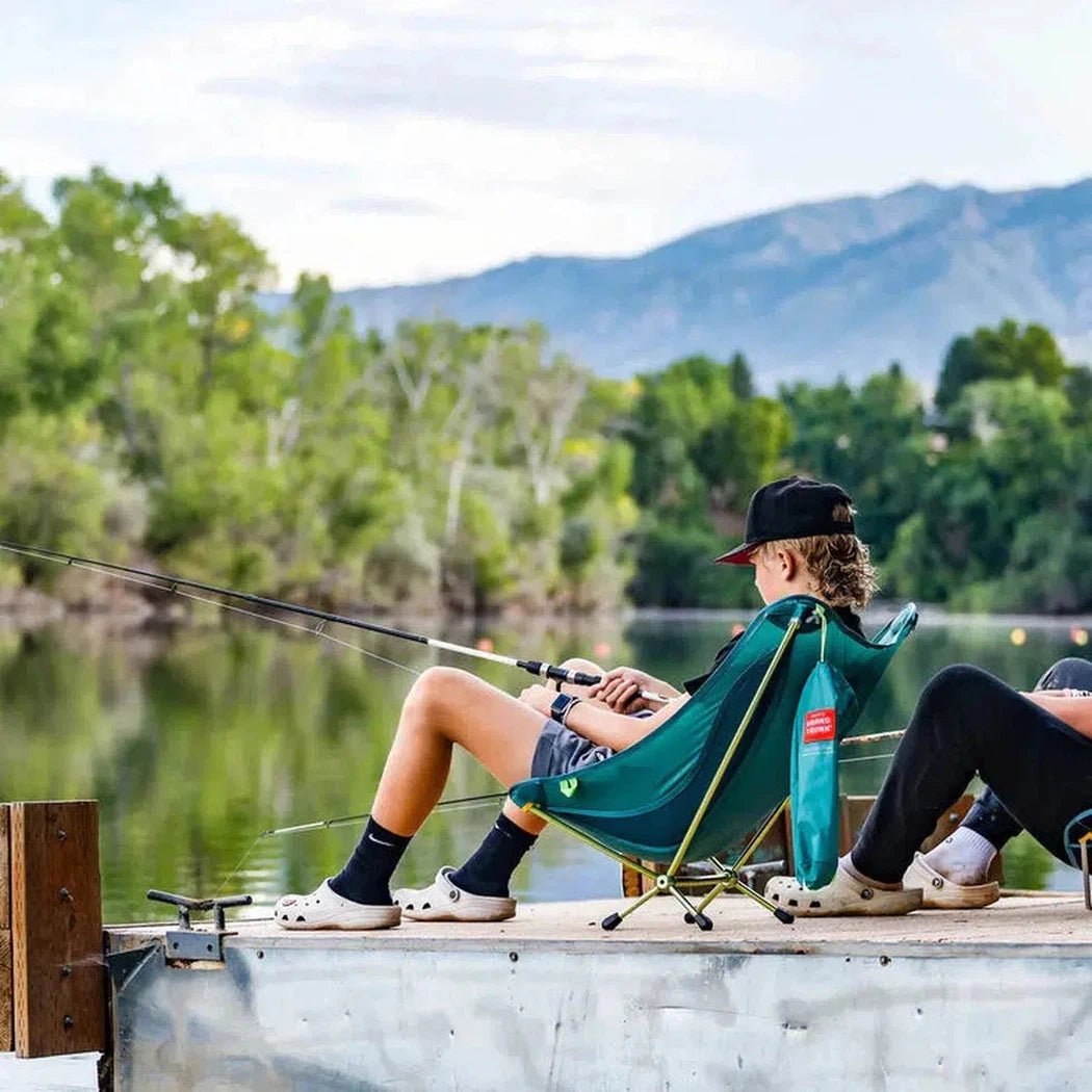 A kid sits in a Black Grand Trunk Mantis chair on a dock by a lake with mountains in the background.