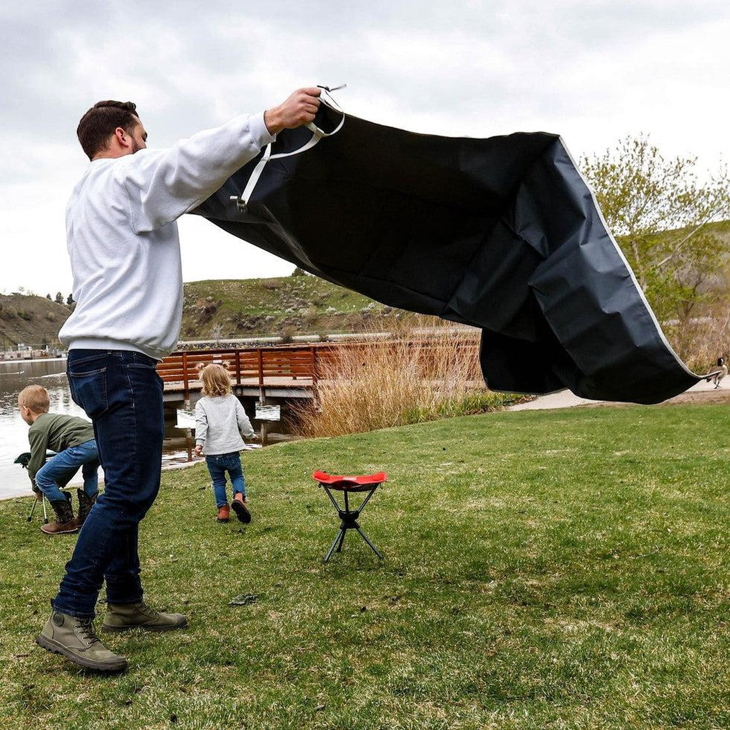 A man holds the Grand Trunk Meadow Mat in Horizon color while two kids play near a lake and a red folding chair sits on grass.