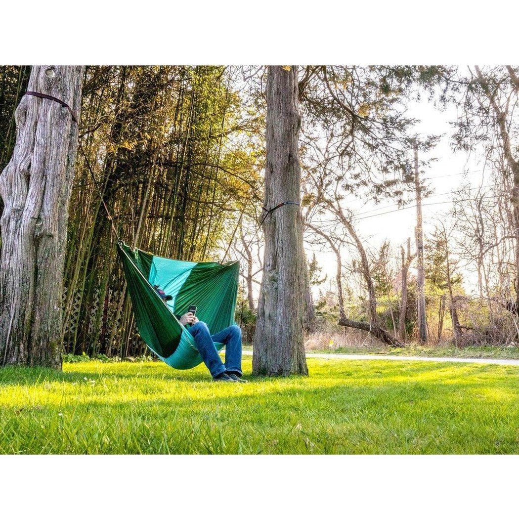 A person relaxes in the Grand Trunk ROVR Hanging Chair in Forest Green/Teal between two trees on a sunny grassy path.