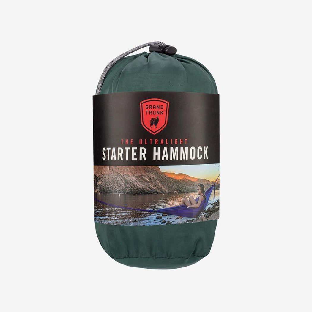 A front view of the Grand Trunk Ultralight Starter Hammock in blue showing the product label and a model relaxing in the hammock against a mountain background.