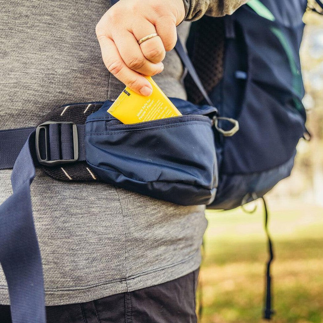 A person puts a yellow card into the pocket of a Gregory Amber 68 Plus backpack in Arctic Navy color, with a backpack strap visible and a grassy background.
