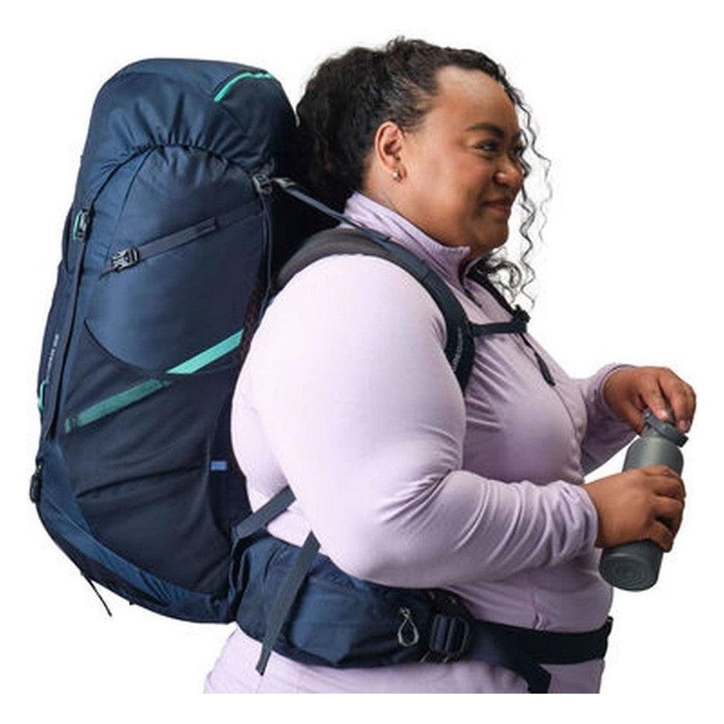 A woman wears the Gregory Amber 68 Plus backpack in Arctic Navy, holding a water bottle against a white background.