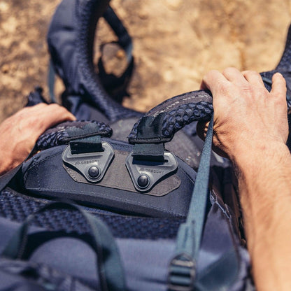 A person’s hands grip the handle of the Gregory Baltoro 65 backpack, which has two metal buckles and a blue strap, against a brown outdoor background.
