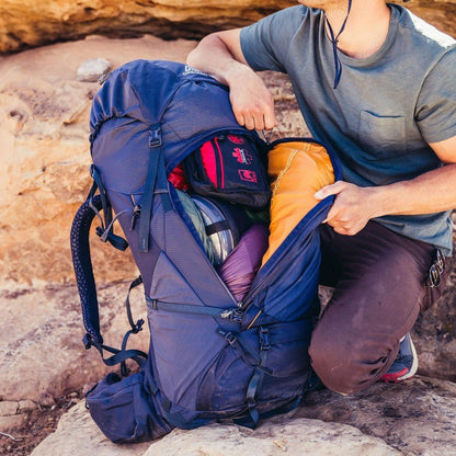 A person kneels by a large blue Gregory Baltoro 65 backpack on rocky terrain, opening its main compartment to show gear inside.