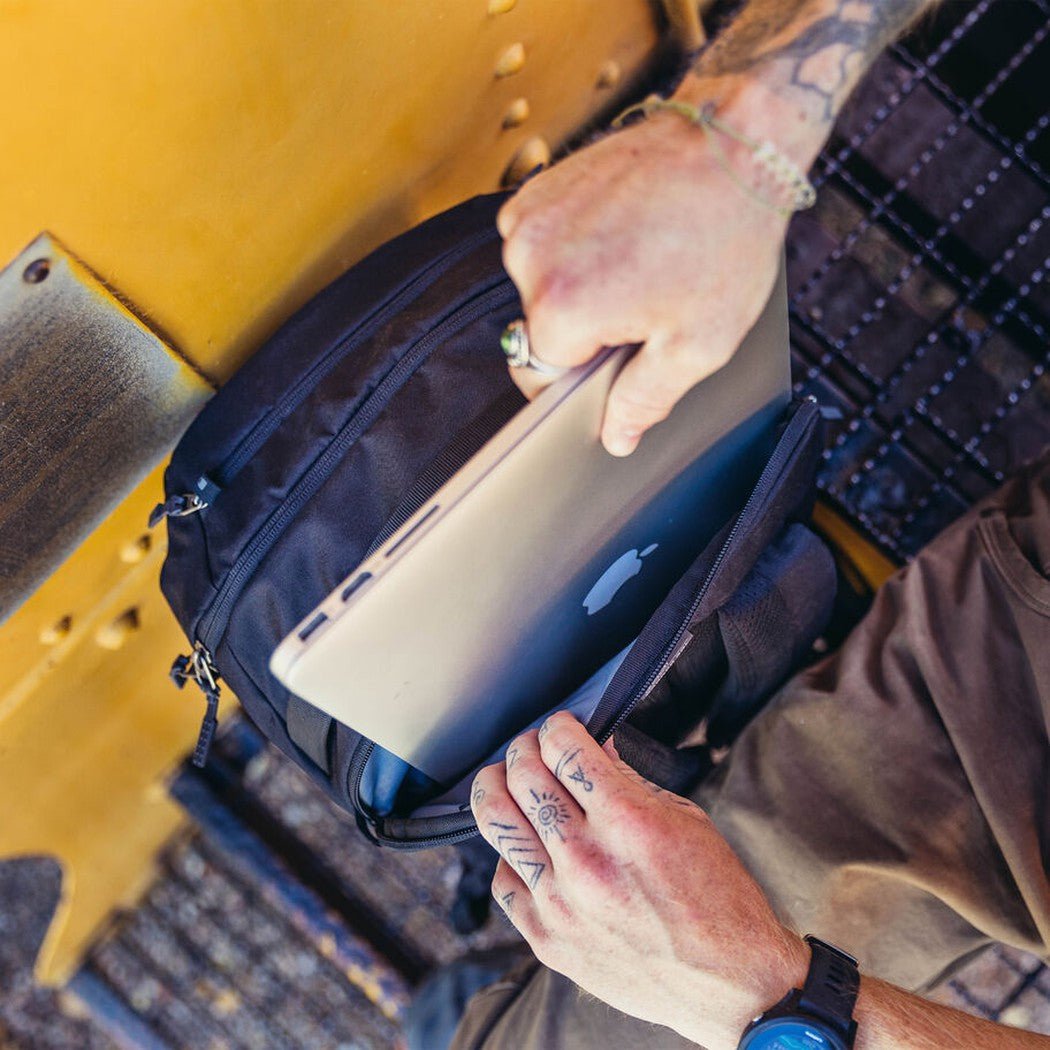 A person puts a silver laptop into the Gregory Border Traveler 30 backpack, which is Total Black, against a yellow metal background.