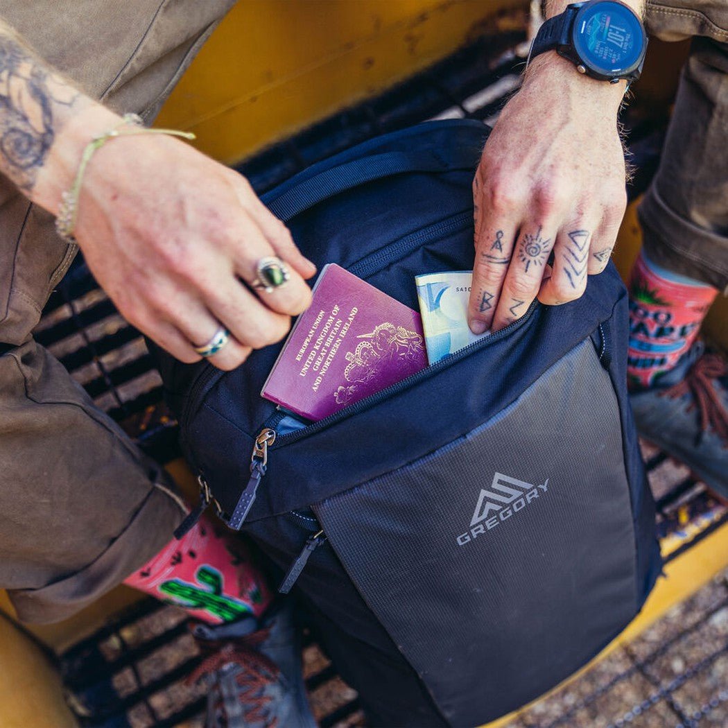 A person puts a passport and cash into the front pocket of a Total Black Gregory Border Traveler 30 backpack.