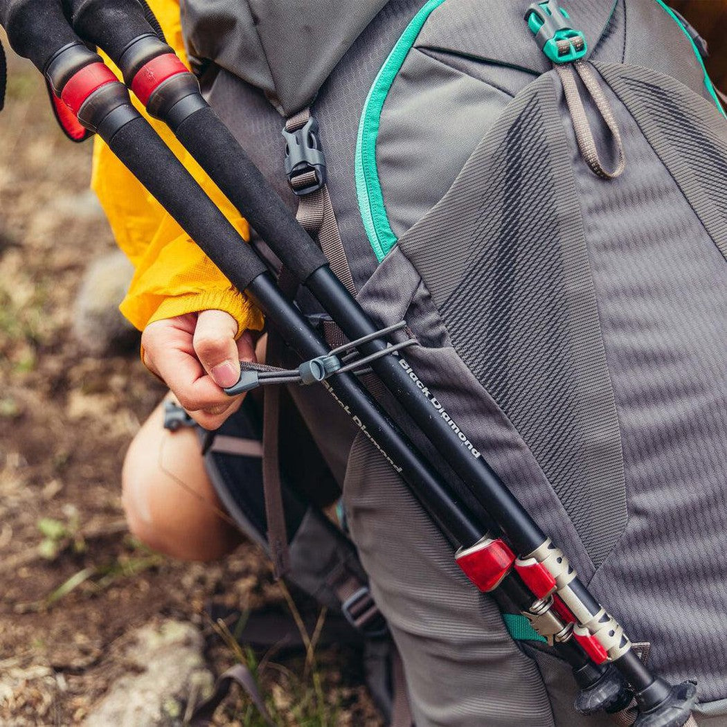 A person in a yellow jacket holds trekking poles attached to a Mist Grey Gregory Jade 43 backpack in a natural outdoor setting.