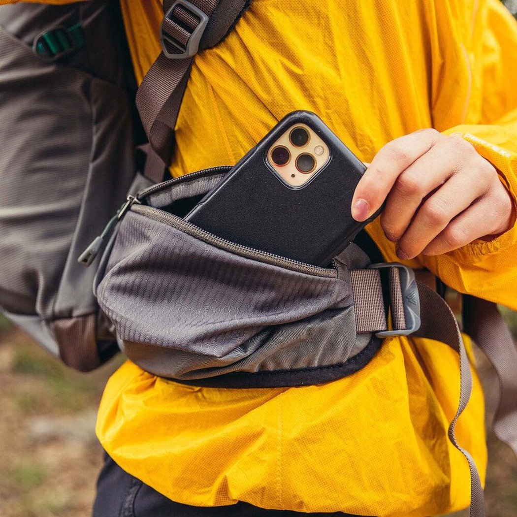 A person in a yellow jacket holds a black phone from the Mist Grey Gregory Jade 43 backpack's front pocket.