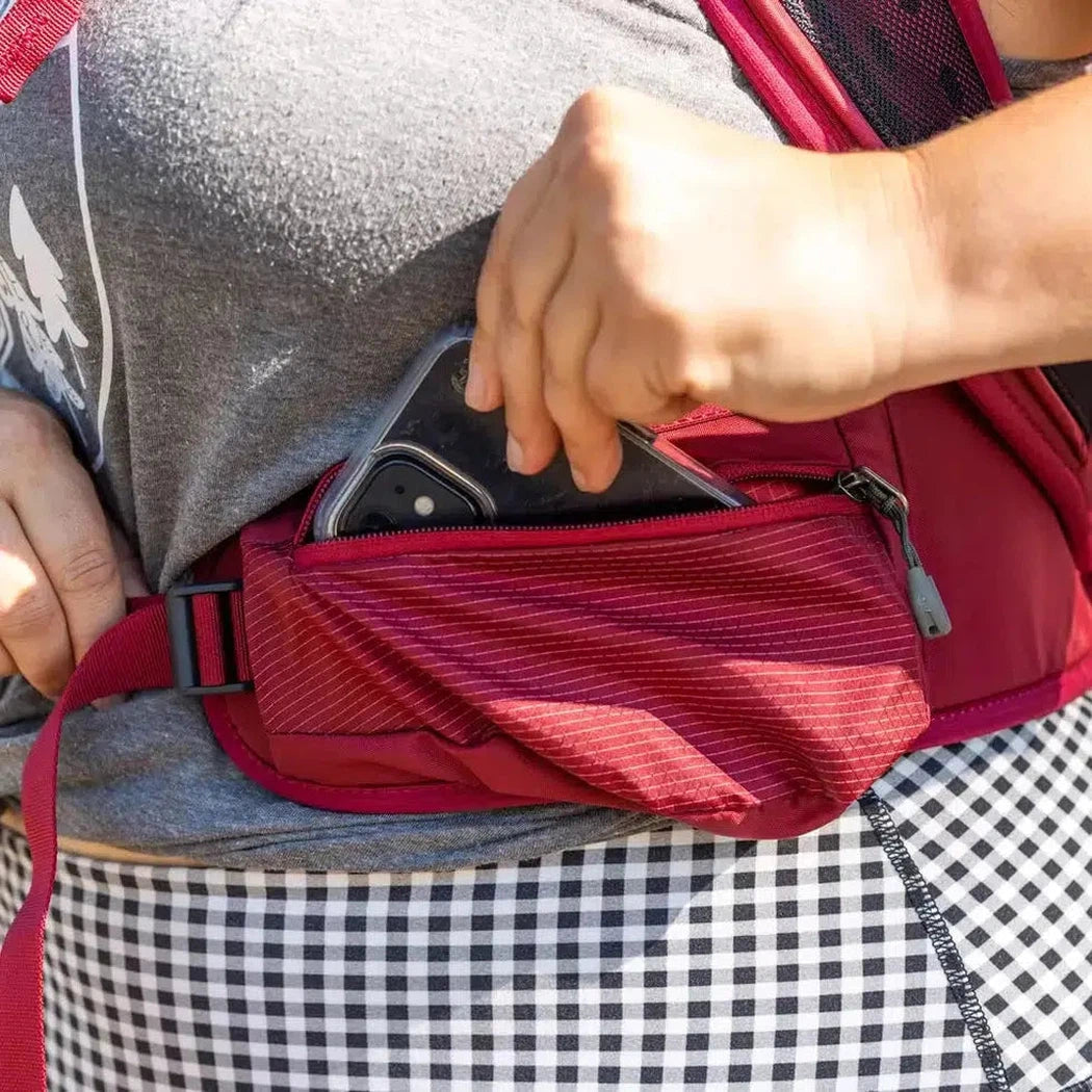 A person wearing a gray shirt and checkered skirt reaches into the Iris Red Gregory Maya 20 Plus daypack's front pocket to grab a phone.