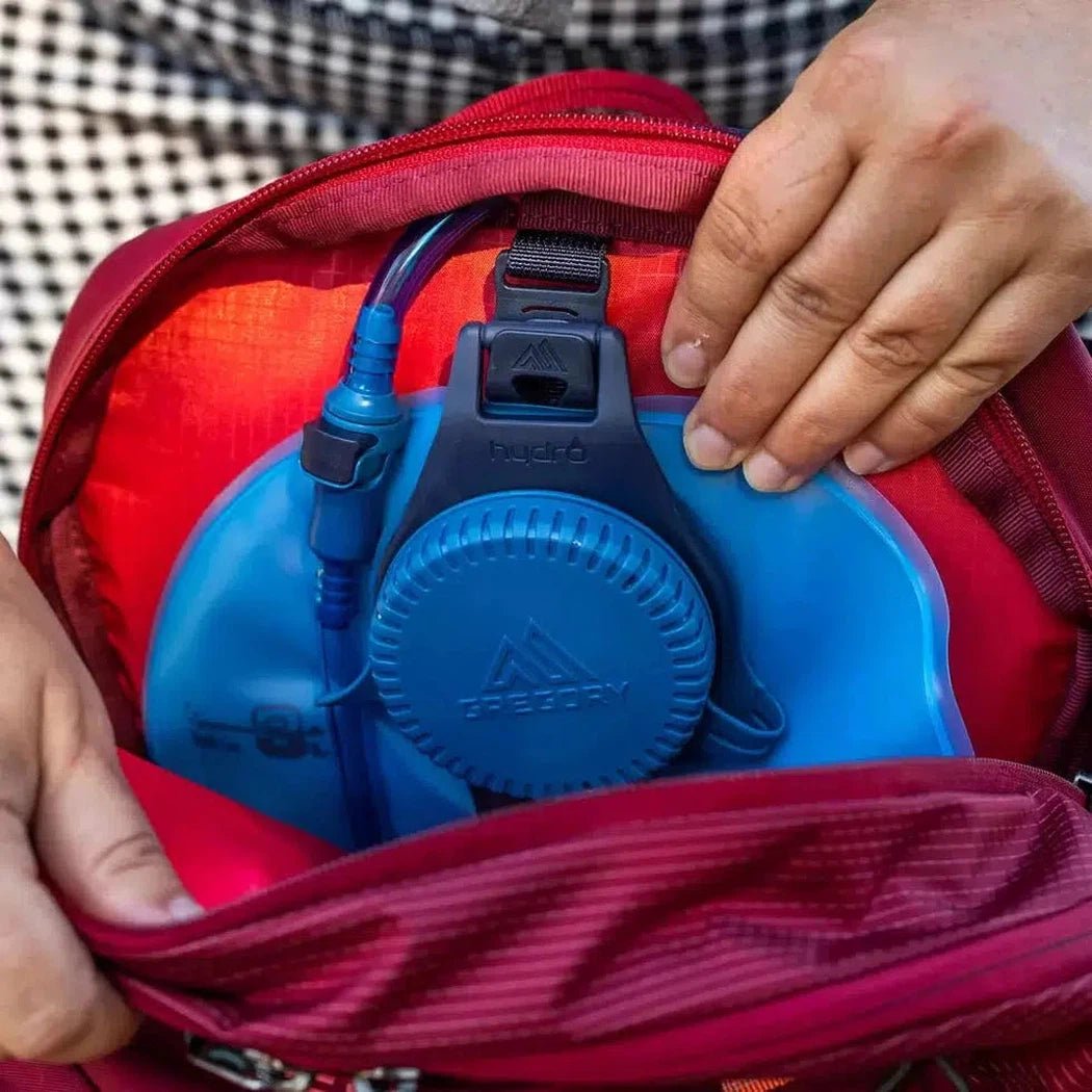 A close-up of the Gregory Maya 20 Plus in Iris Red showing the blue water reservoir and a person's hand on the backpack.