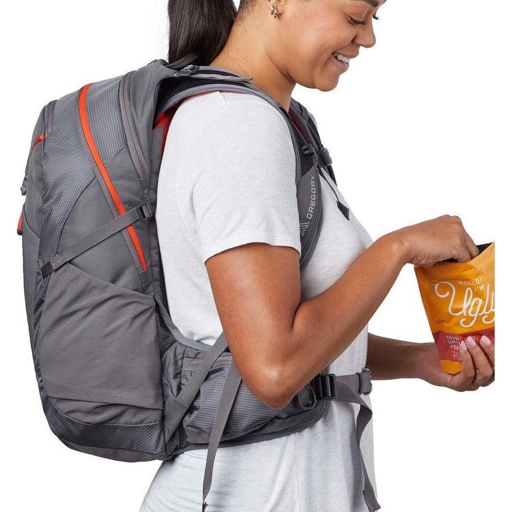 A woman in a white tee wears the Gregory Maya 20 backpack with a side pocket visible, holding a snack bag against a white background.