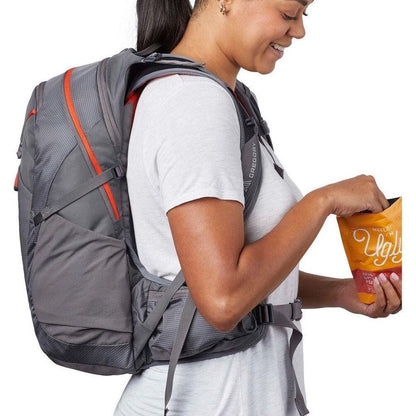 A woman in a white tee wears the Gregory Maya 20 backpack with a side pocket visible, holding a snack bag against a white background.