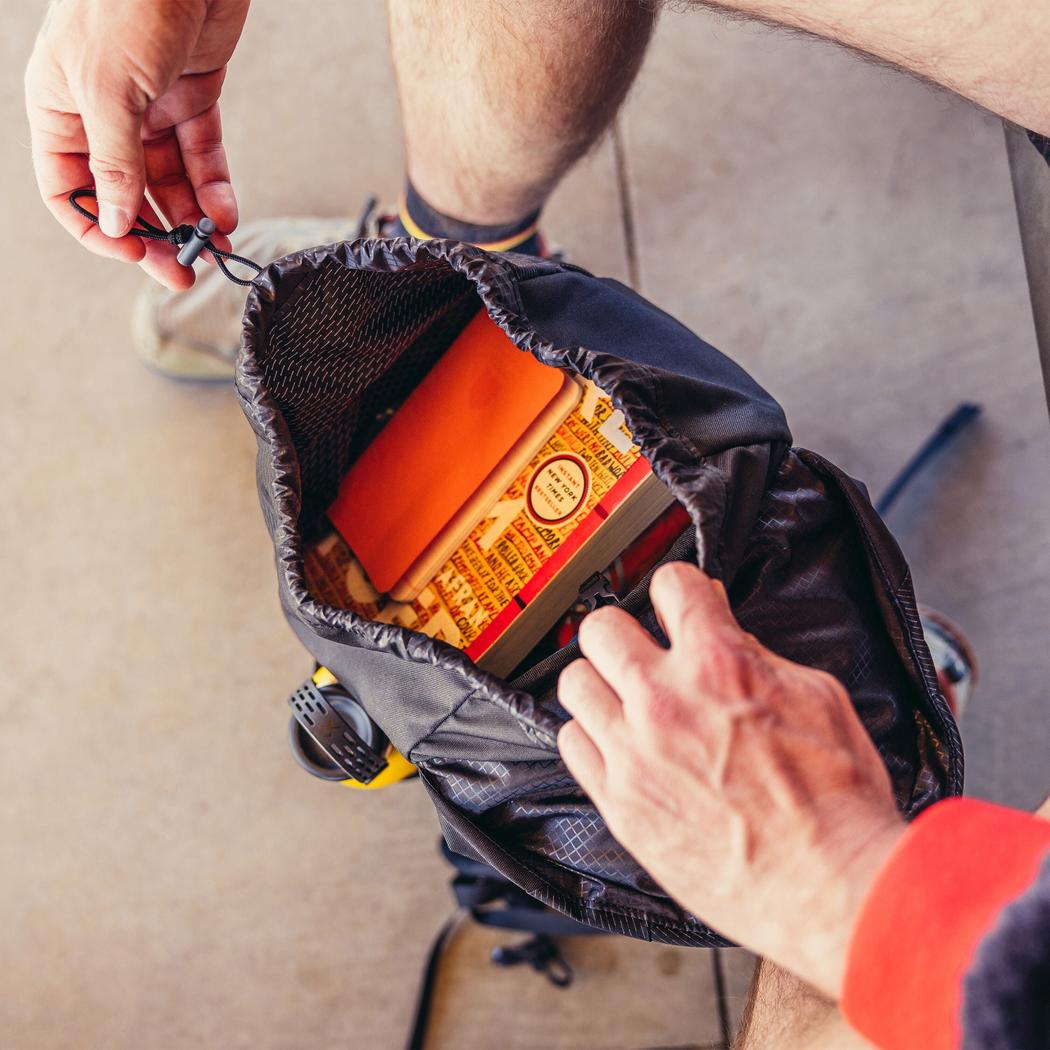 A person opens the Gregory Nano 16 daypack, showing its interior with a Moab Tan box and a camera strap visible.