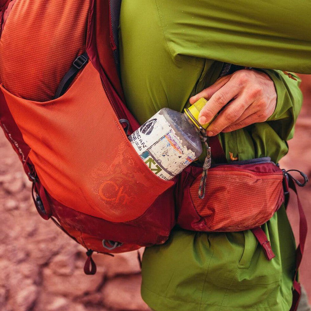 A close-up of a person wearing the Gregory Paragon 58 Ferrous Orange backpack with a water bottle in the side pocket, set against a rocky outdoor background.