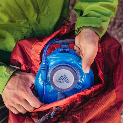 A person in a green jacket holds a blue Gregory Paragon 58 water bladder inside a red Ferrous Orange backpack.