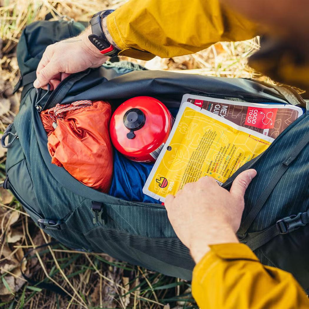 A person in a yellow jacket opens the Gregory Paragon 70 backpack, revealing a red kettle, orange stuff sack, and yellow food packet inside, set against a grassy background.