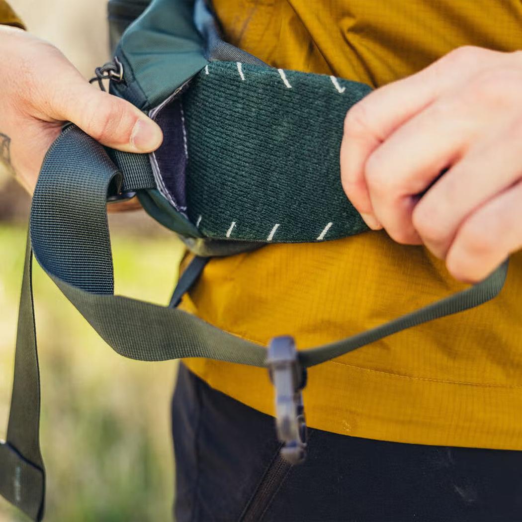 A person adjusts the Gregory Paragon 70 backpack's shoulder strap in Cascade Green against a blurred outdoor background.