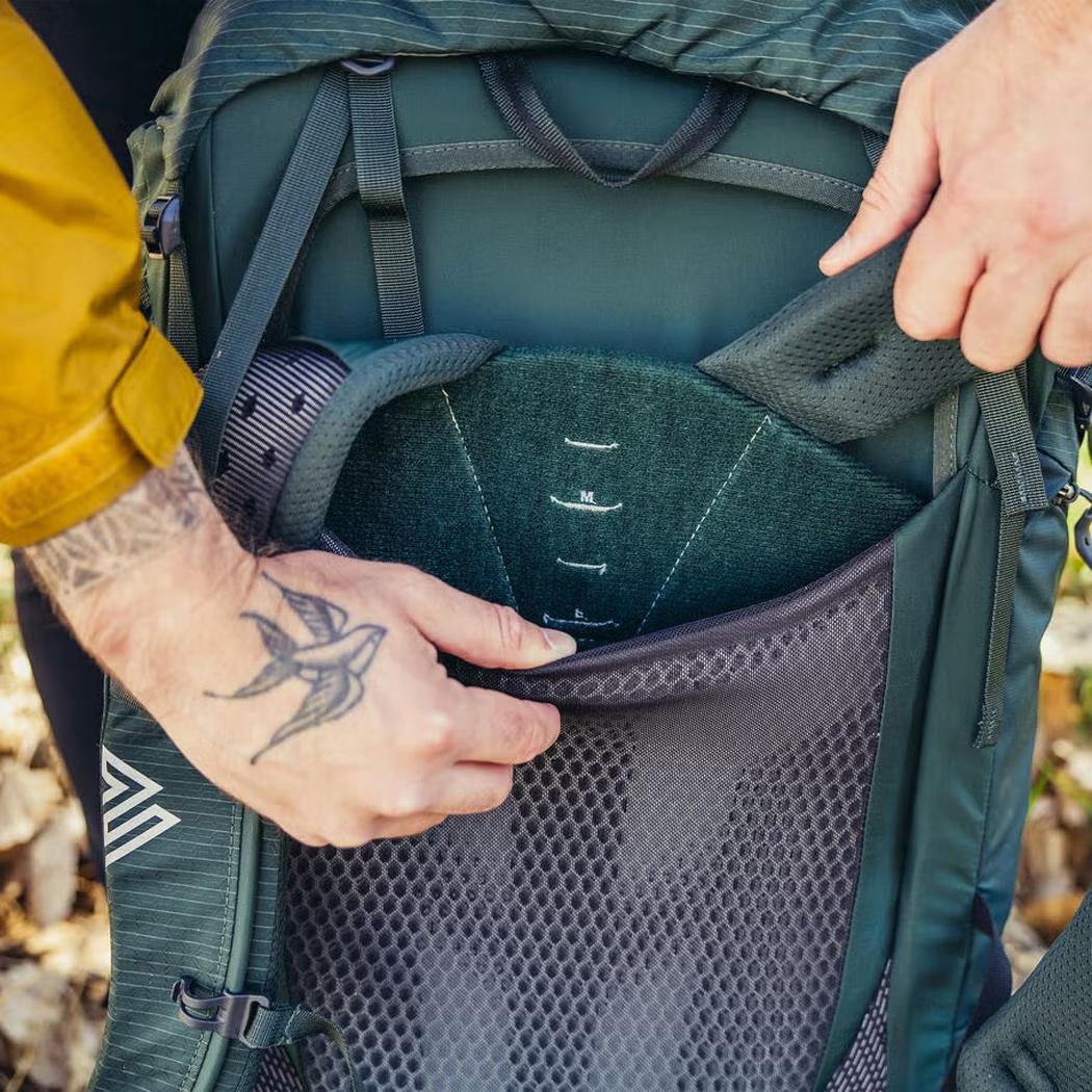 A person with a tattooed hand adjusts the Gregory Paragon 70 backpack's front pocket, showing its Cascade Green color and mesh panel.
