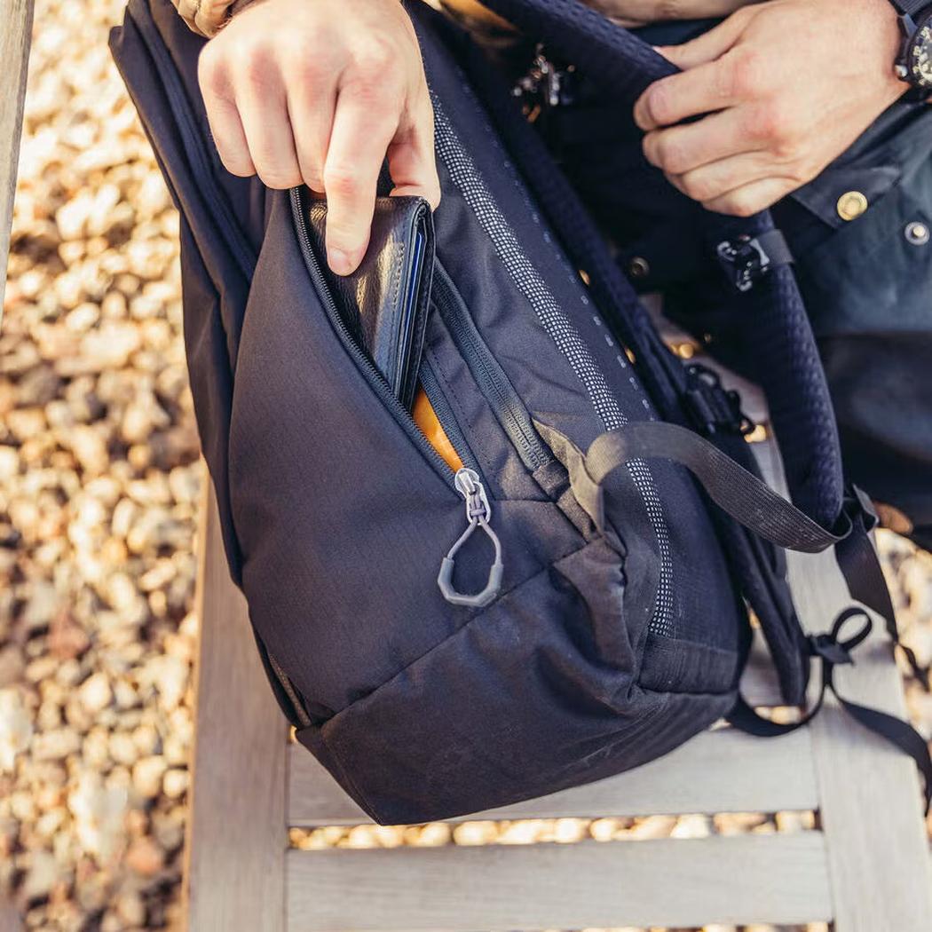 A person reaches into the front pocket of the Gregory Rhune 22 daypack, which is Cabon Black, while sitting on a chair with a gravel background.