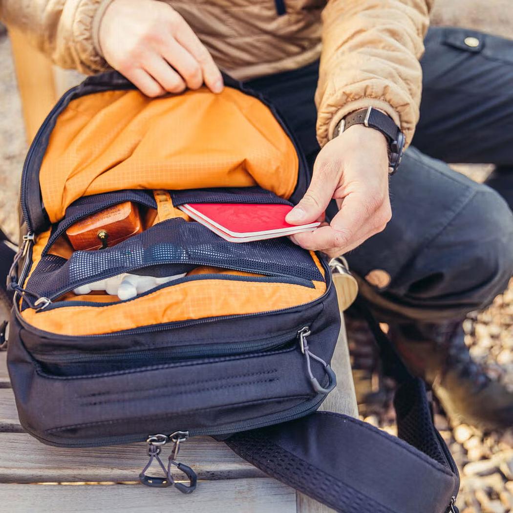 A person puts a red phone in the Gregory Rhune 22 daypack, which is Cabon Black with an orange interior, on a wooden bench.