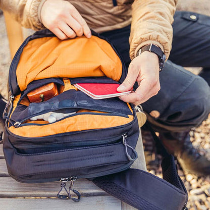 A person puts a red phone in the Gregory Rhune 22 daypack, which is Cabon Black with an orange interior, on a wooden bench.