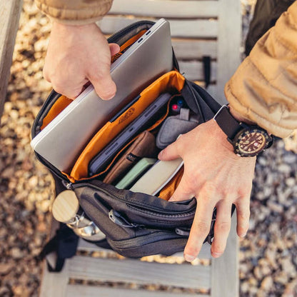 A person wearing a watch puts a laptop into the Gregory Rhune 22 daypack, which is Cabon Black and has an orange interior, on a wooden bench with gravel.
