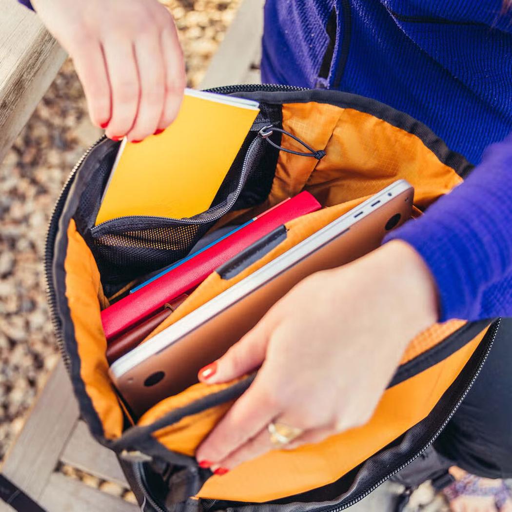 A person in a blue long-sleeve top reaches into the open Gregory Rhune 25 daypack, which has a bright orange interior and holds a tablet, a red notebook, and a yellow book, with a gravel background.
