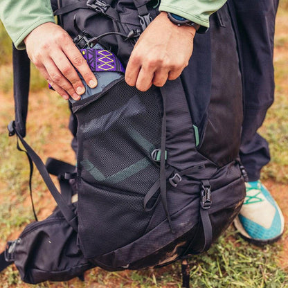 A person puts a purple item in the front pocket of a Forest Black Gregory Stout 70 backpack on grass.