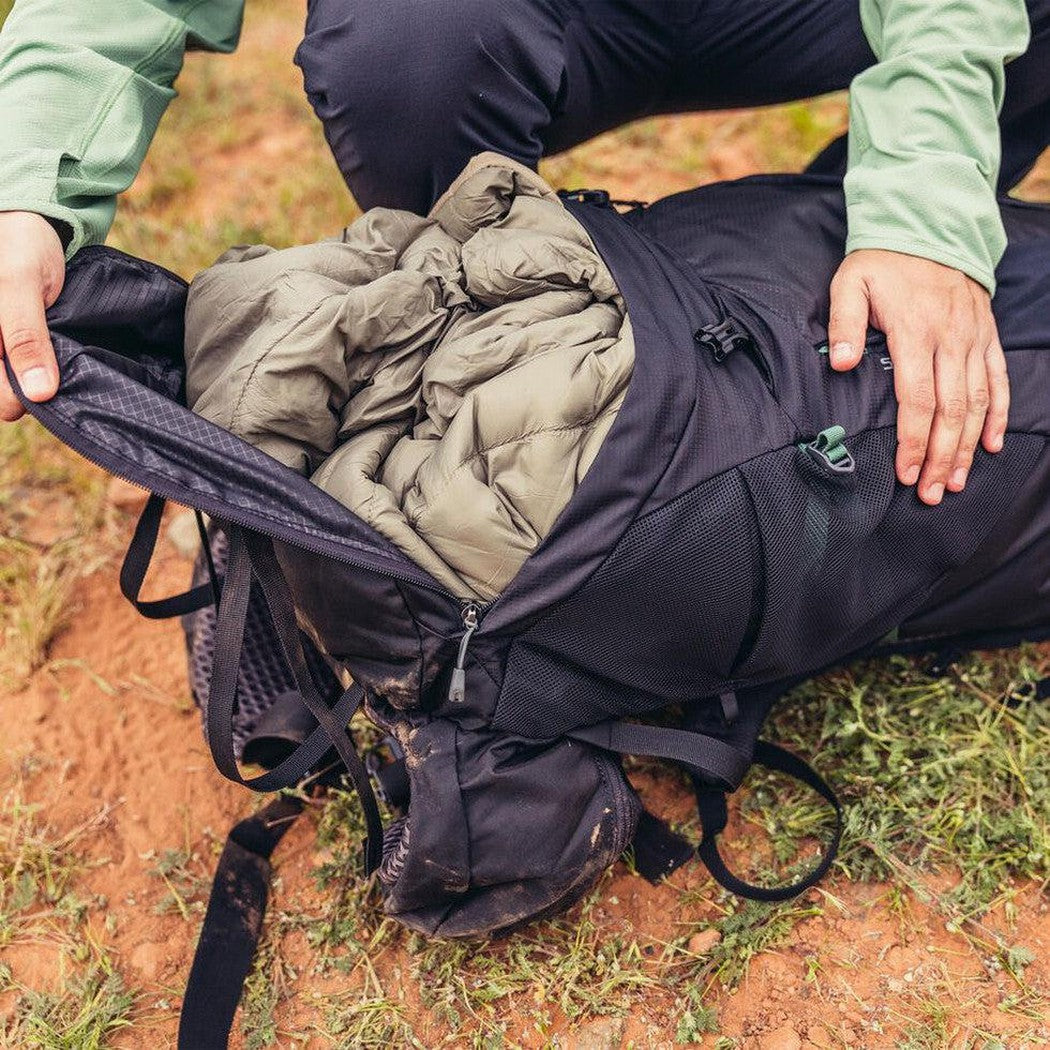 A person crouches on dirt and grass, opening the Forest Black Gregory Stout 70 backpack to show its interior.