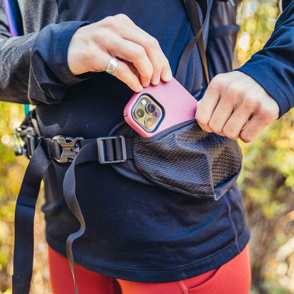 A person puts a pink phone into the Antigua Green Gregory Sula 8 H20 daypack's front pocket in an outdoor setting.