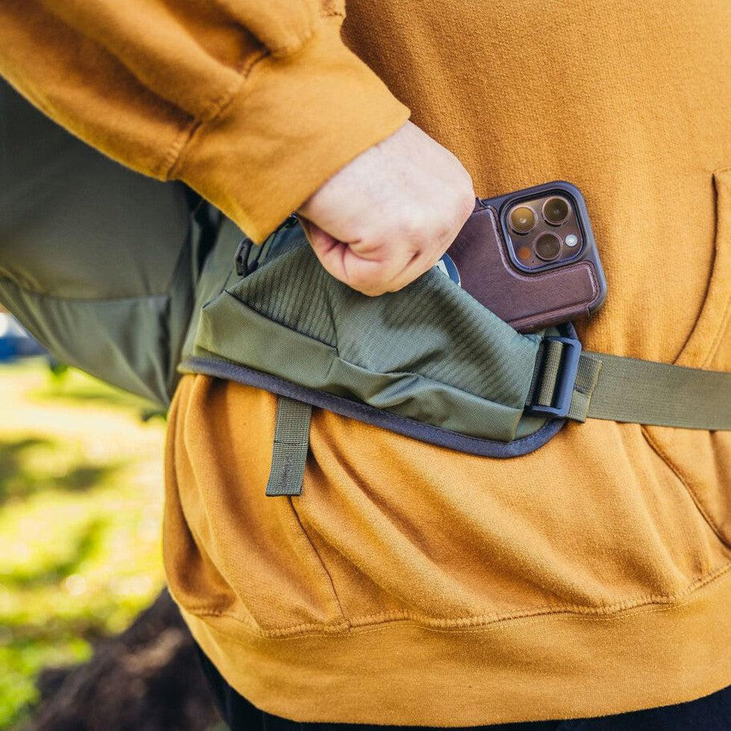 A person in a mustard yellow sweatshirt wears the Gregory Zulu 28 LT Plus in Forage Green, with a phone in the side pocket, against a blurred outdoor background.