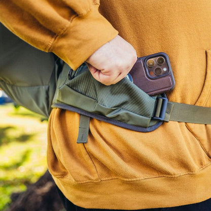 A person in a mustard yellow sweatshirt wears the Gregory Zulu 28 LT Plus in Forage Green, with a phone in the side pocket, against a blurred outdoor background.