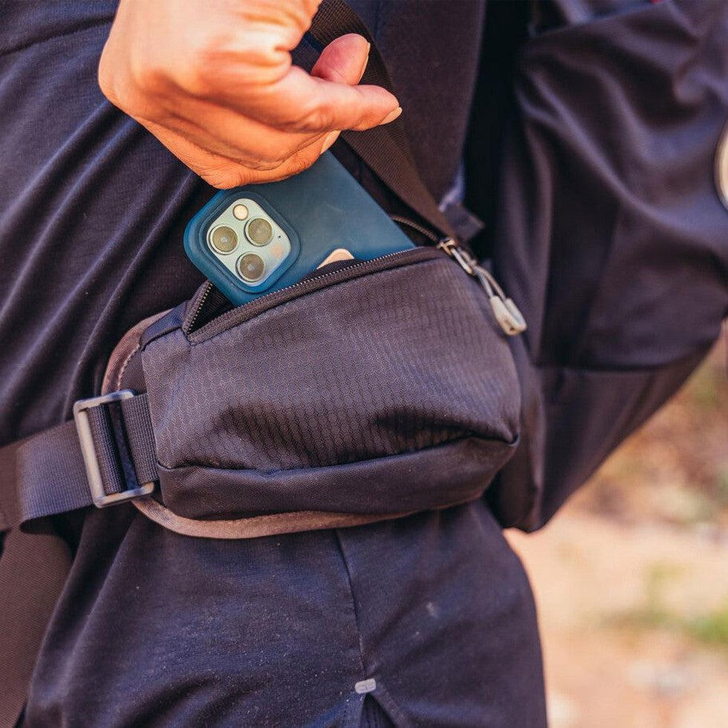 A person wearing the Gregory Zulu 28 LT Forage Green daypack reaches into the front pocket to grab a blue phone, with a blurred outdoor background.