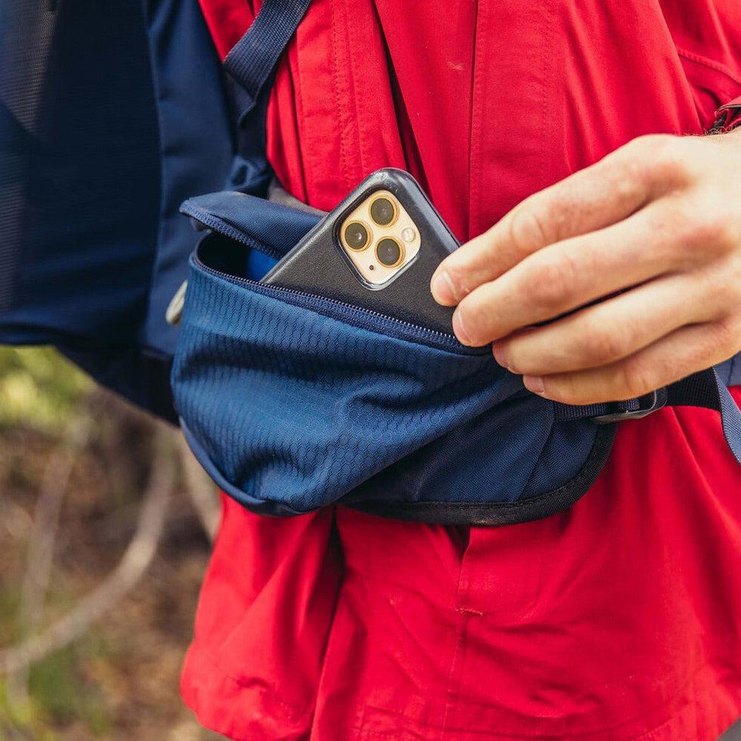 A person in a red jacket reaches into a blue pocket of the Gregory Zulu 45 daypack to grab a phone, with a forest background.