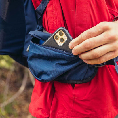 A person in a red jacket reaches into a blue pocket of the Gregory Zulu 45 daypack to grab a phone, with a forest background.