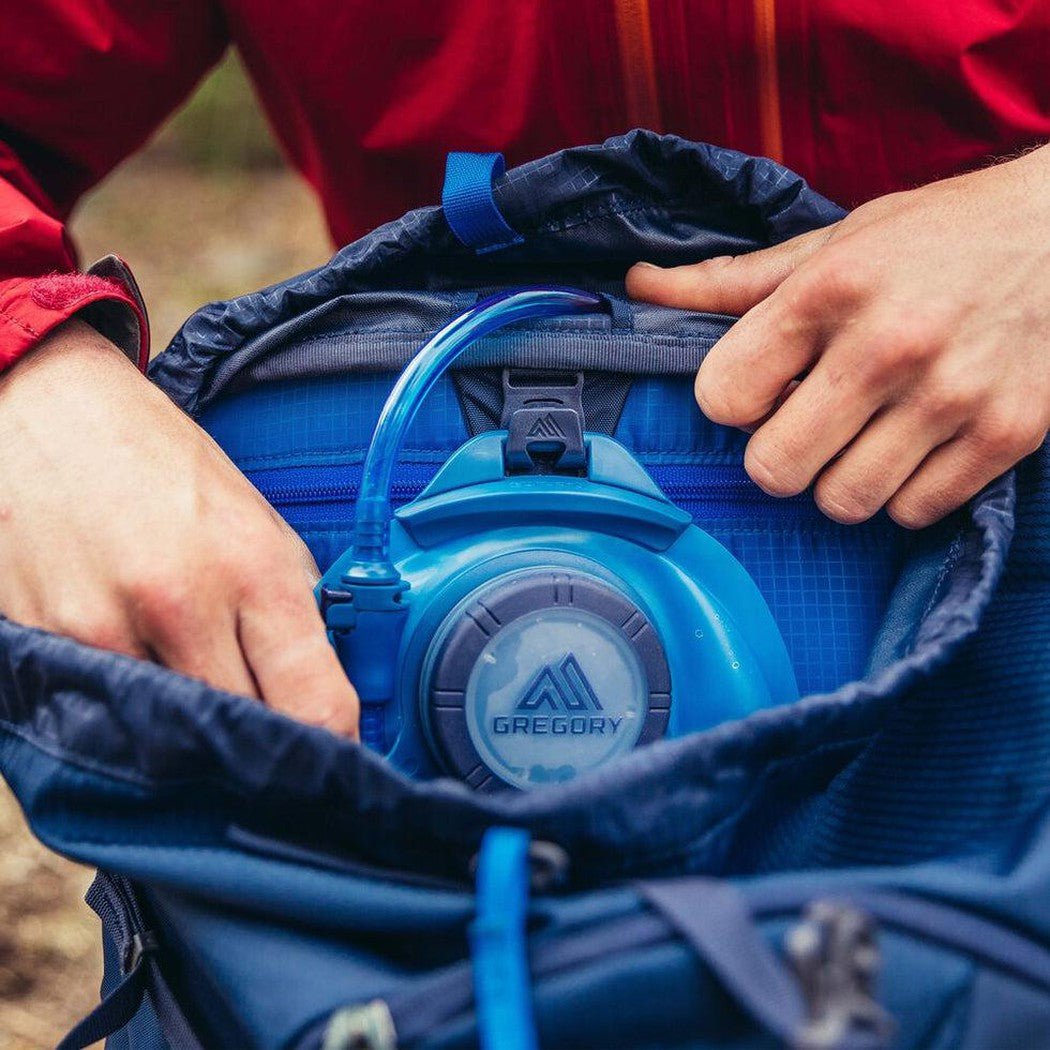 A person in a red jacket opens a blue Gregory Zulu 45 daypack to show its interior and a blue Gregory water reservoir.
