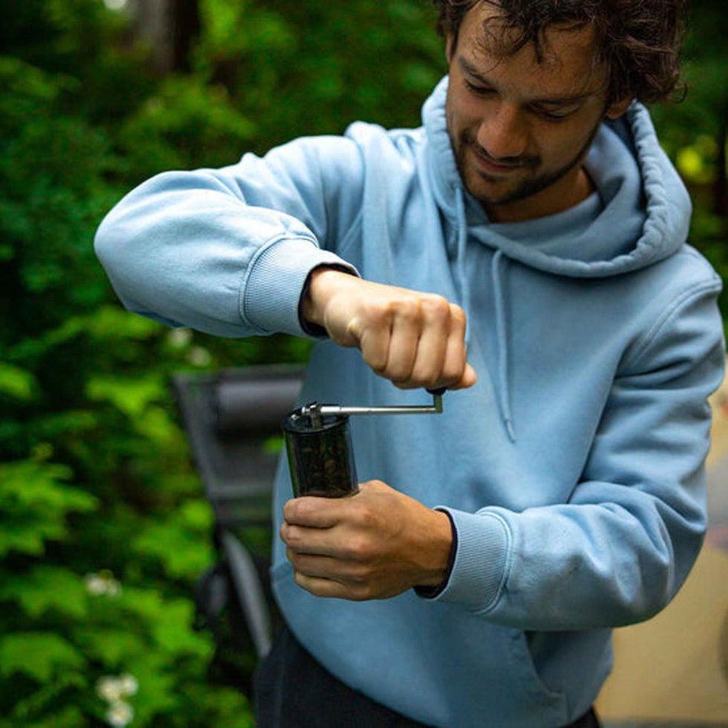 A man in a light blue hoodie grinds coffee with the GSI Outdoors Javagrind in a lush green outdoor setting.