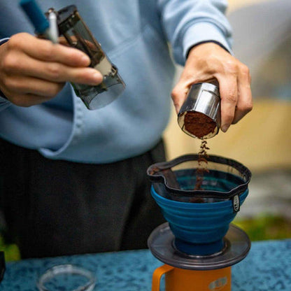 A person pours ground coffee from a GSI Outdoors Javagrind into a blue collapsible coffee filter over an orange camping coffee maker on a blue table.