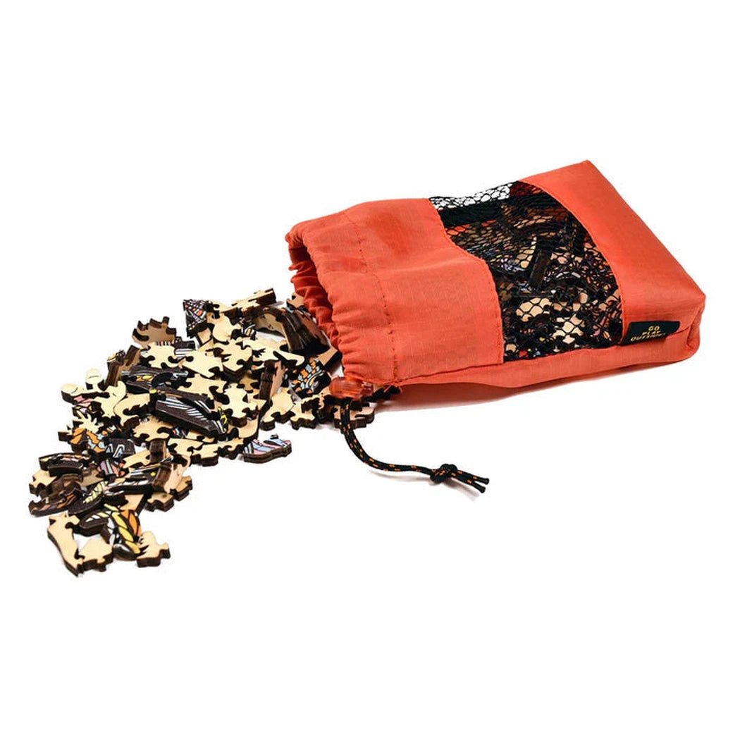 An orange GSI Outdoors Wood Puzzle bag with a black mesh panel lies on its side spilling puzzle pieces onto a white background.