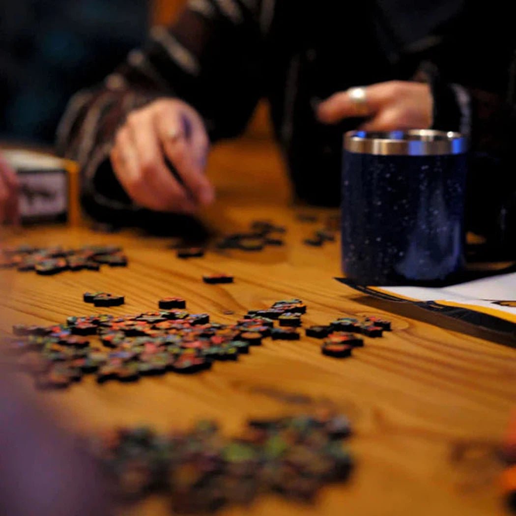 A person works on a GSI Outdoors Wood Puzzle on a wooden table with a blue cup nearby.
