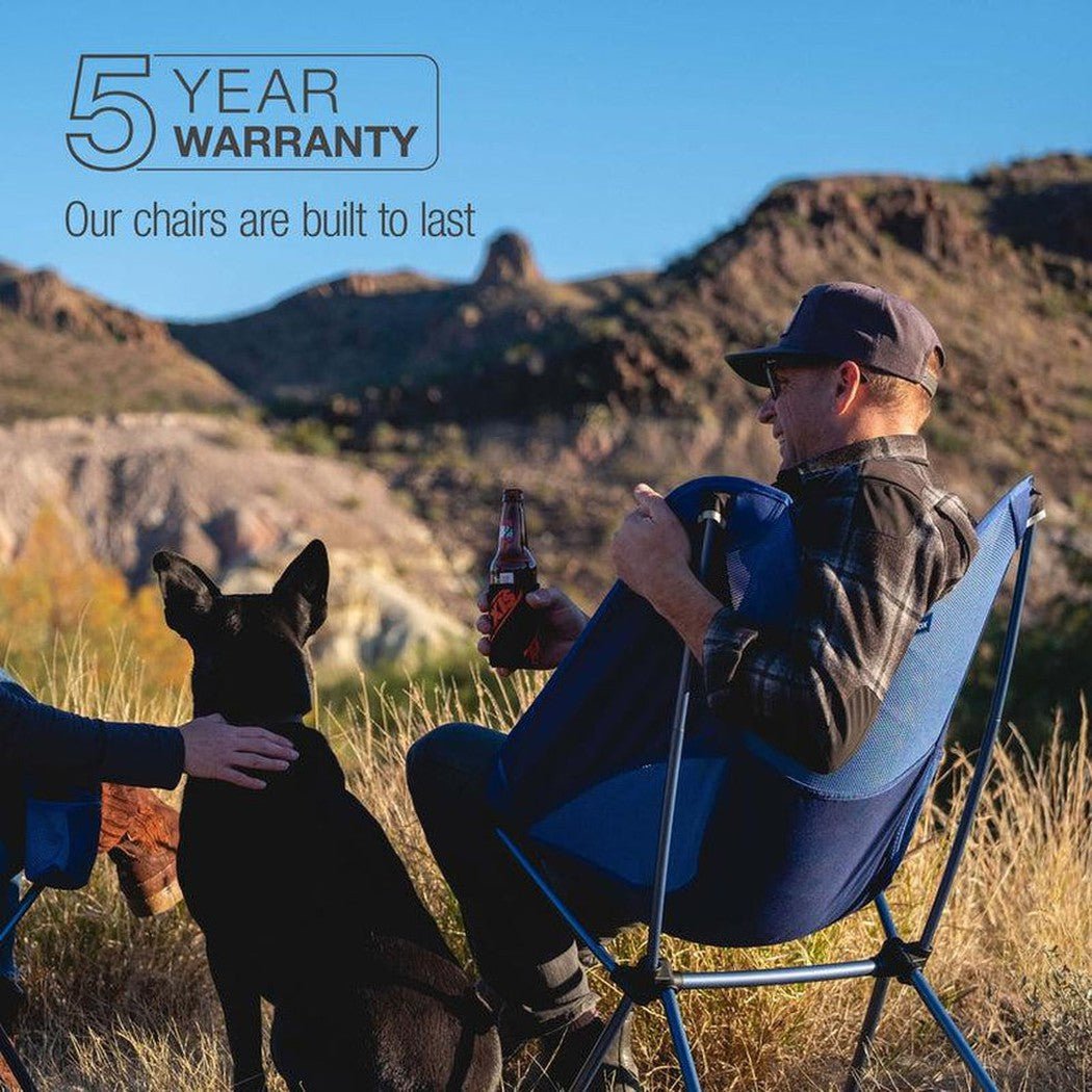A man sits in a Black Helinox Chair One XL with a dog nearby, set against a mountainous outdoor backdrop.