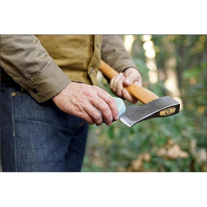 A person in a brown shirt and blue jeans holds the Helko Werk Axe Sharpening Stone against the axe blade, with a forest background.