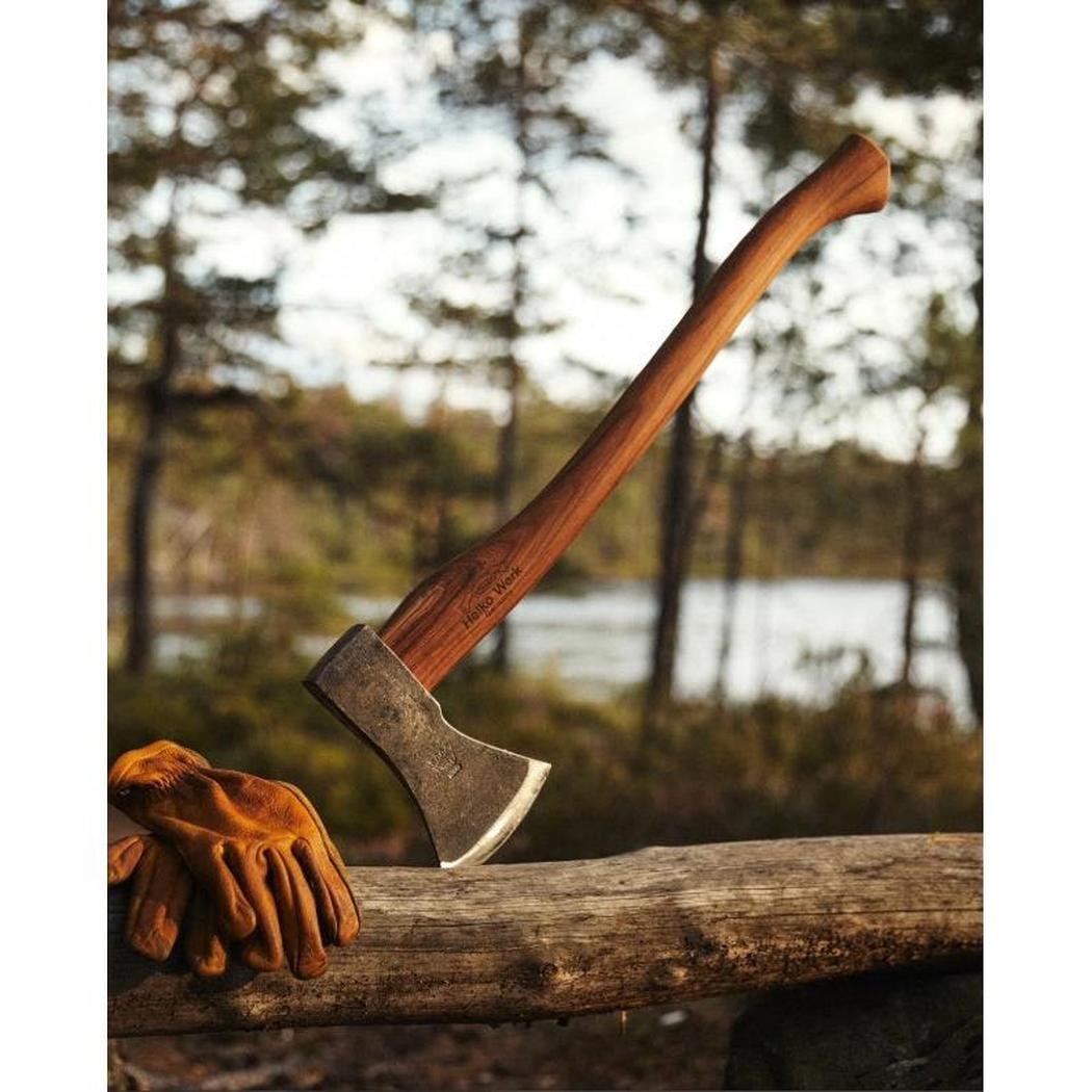 A Helko Werk Black Forest Woodworker axe rests on a log with brown gloves, set against a forest and lake background.