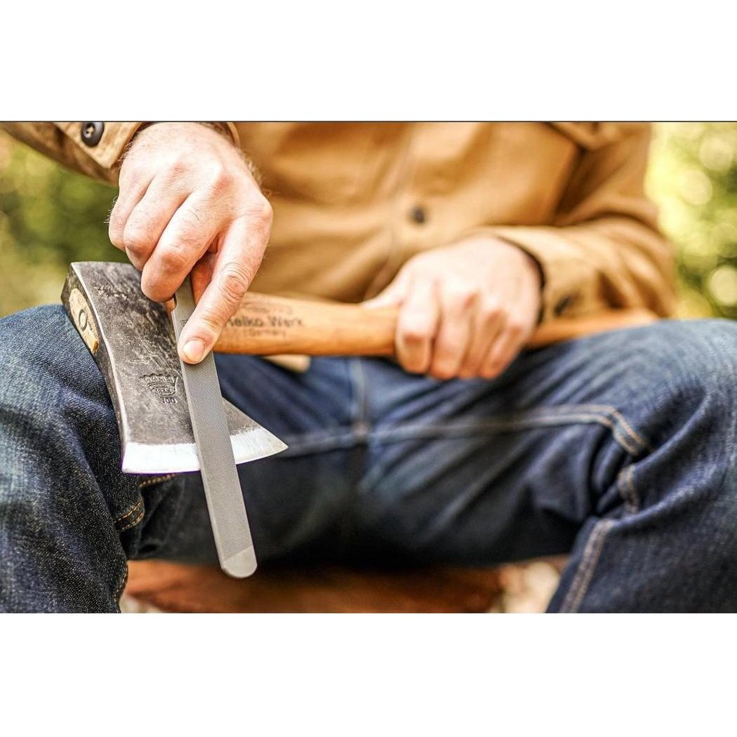 A person in blue jeans sharpens a Helko Werk Compact Sharpening File on an axe with a wooden handle, outdoors.