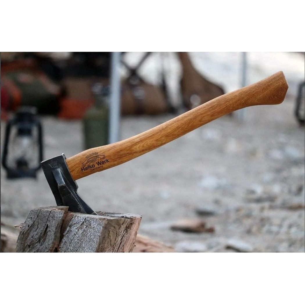 A close-up of the Helko Werk Heidelberg Mini Spalter axe with a wooden handle resting on a log, showing the brand name and a camping background.