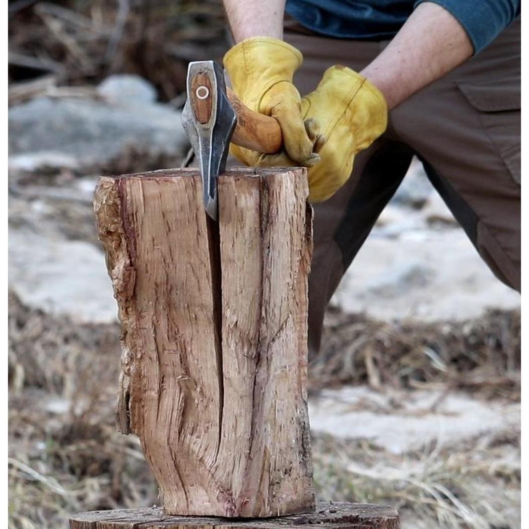 A person in yellow gloves holds the Helko Werk Heidelberg Mini Spalter axe on a wooden stump outdoors.