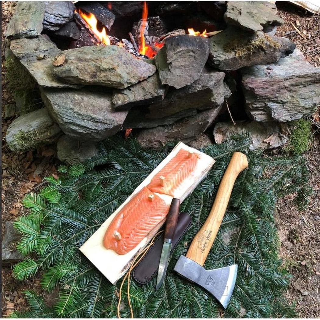A Helko Werk Rheinlander Hatchet rests on evergreen foliage next to a piece of salmon and a knife, with a campfire in the background.