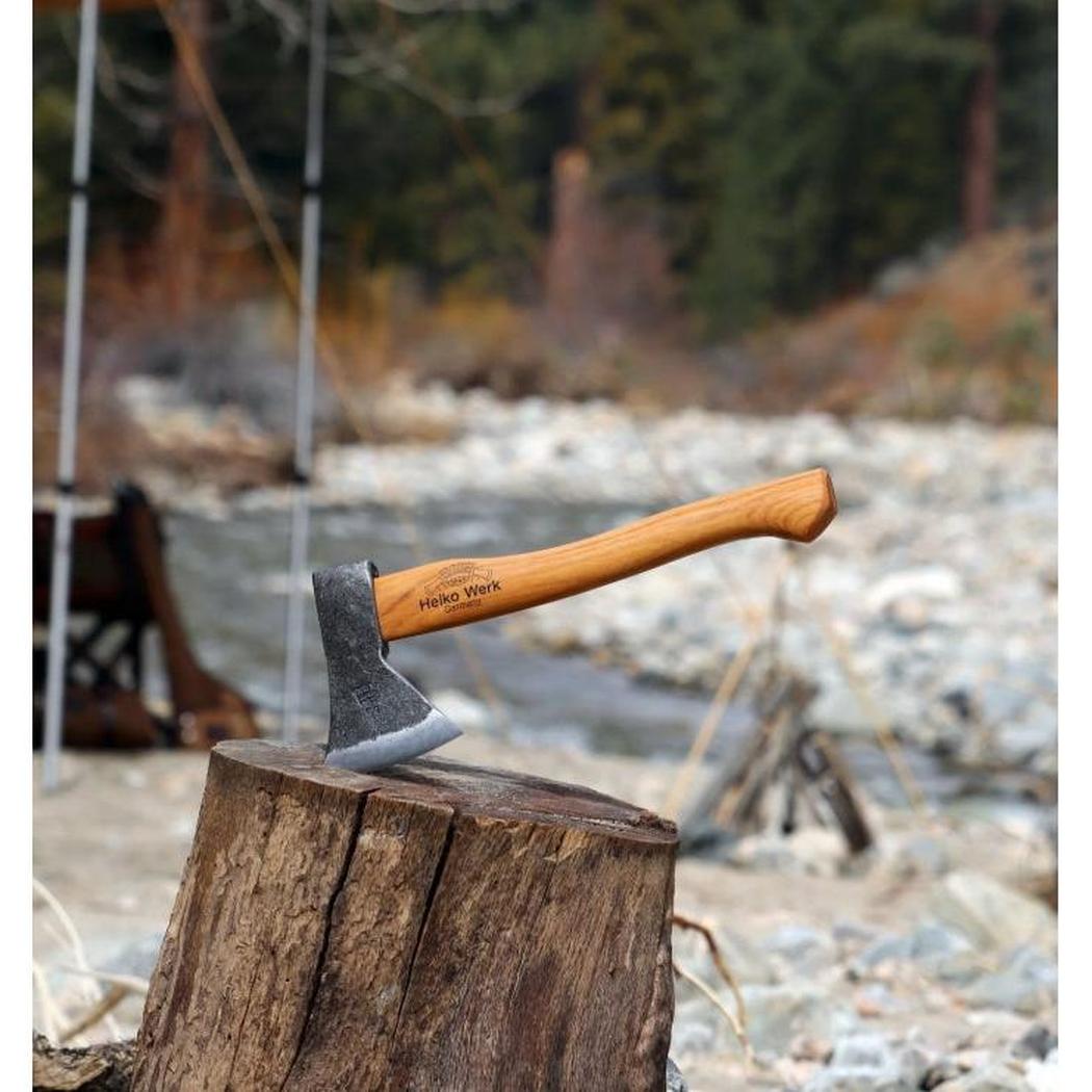 A Helko Werk Rheinlander Hatchet rests on a tree stump with a river and campsite in the background.