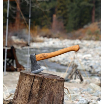 A Helko Werk Rheinlander Hatchet rests on a tree stump with a river and campsite in the background.